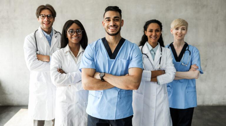 International team of doctors posing together while having morning breefing, male and female medical workers standing against wall, holding arms crossed on chests, cheerfully smiling at camera